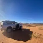 Silver SUV parked on sand dunes with mountains in the background under a clear blue sky.